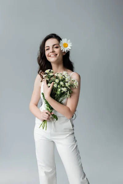 Positive woman in white clothes holding bouquet of chamomiles isolated on grey — Stock Photo