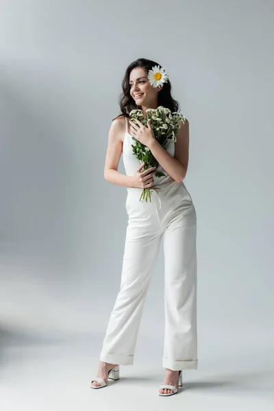 Full length of trendy woman in white clothes and heels holding chamomiles on grey background — Fotografia de Stock