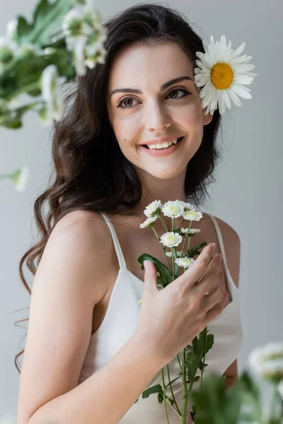 Smiling woman in white top looking at blurred chamomiles isolated on grey — Stockfoto