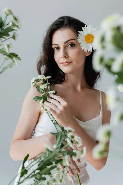 Brunette woman in white top holding chamomiles and looking at camera isolated on grey — Photo de stock