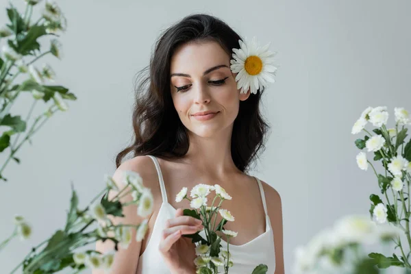 Smiling woman in white top touching flowers isolated on grey - foto de stock