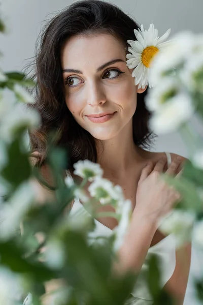 Усміхнена жінка з вірусом і ромашкою в волоссі дивиться далеко біля розмитих рослин ізольовані на сірому — стокове фото