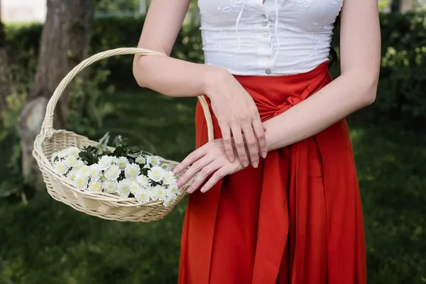 Cropped view of woman in blouse and skirt holding wicker basket with flowers in park — Foto stock