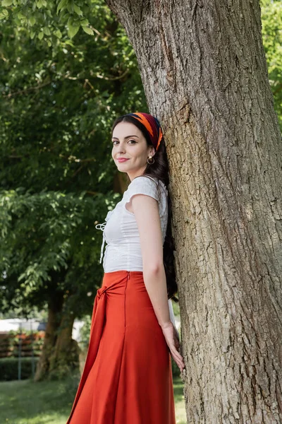 Красивая модель в блузке и юбке, стоящая возле дерева в парке — стоковое фото
