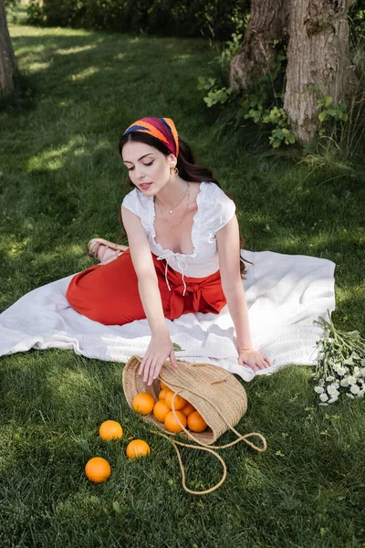 Молодая модная женщина смотрит на апельсины в мешке рядом с цветами в парке — стоковое фото