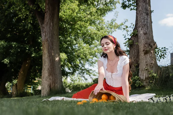 Stylish woman looking at oranges in straw bag on blanket in park - foto de stock