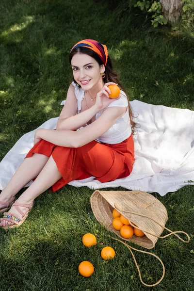 High angle view of cheerful woman holding orange on blanket in park - foto de stock