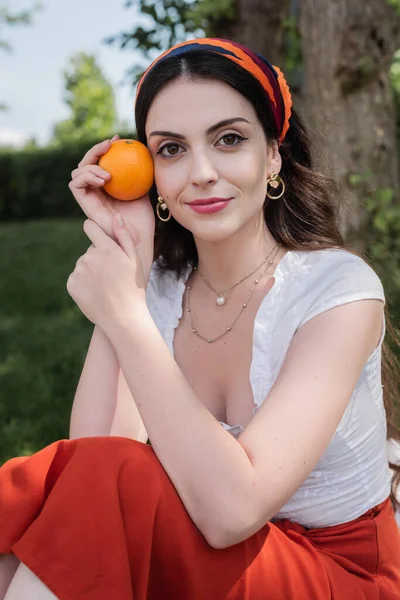 Young woman in blouse and skirt holding orange in park - foto de stock