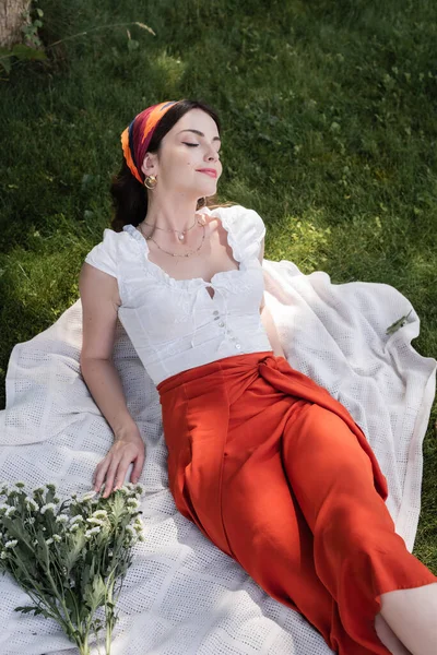 High angle view of stylish woman in blouse resting on blanket near flowers in park — Foto stock