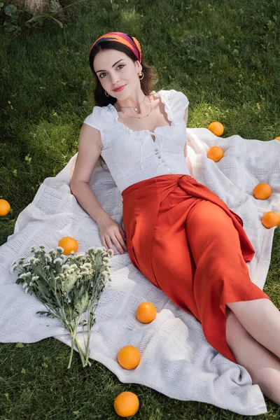 High angle view of stylish woman looking at camera near flowers and oranges on blanket in park — Photo de stock