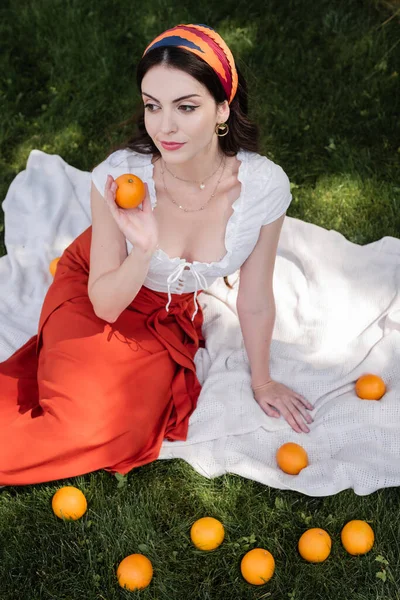 Stylish woman holding orange while sitting on blanket on grass — Photo de stock