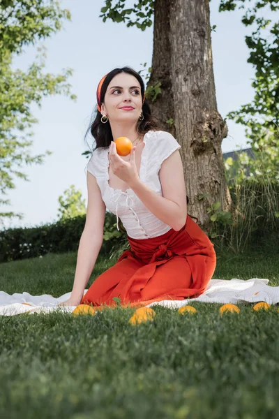 Brunette woman in blouse and skirt holding oranges on blanket in park — Stock Photo