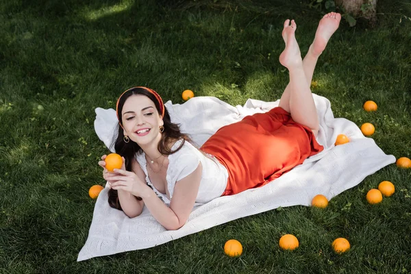 Cheerful barefoot woman in blouse holding orange while lying on blanket in park - foto de stock