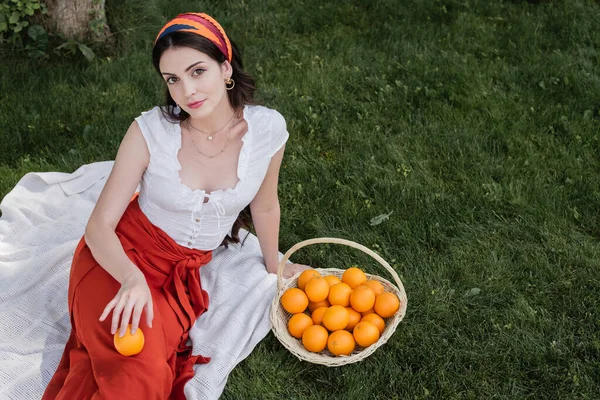 High angle view of smiling woman in blouse holding orange while sitting on blanket in park - foto de stock