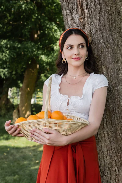 Smiling woman in blouse holding wicker basket with oranges in park — Fotografia de Stock