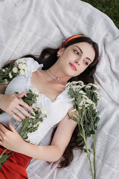 Top view of smiling woman holding daisies while lying on blanket in park - foto de stock