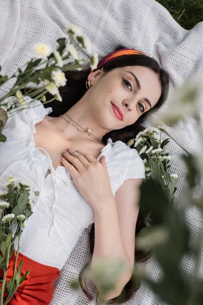 Top view of pretty woman in blouse looking at camera near chamomiles on blanket in park — Fotografia de Stock