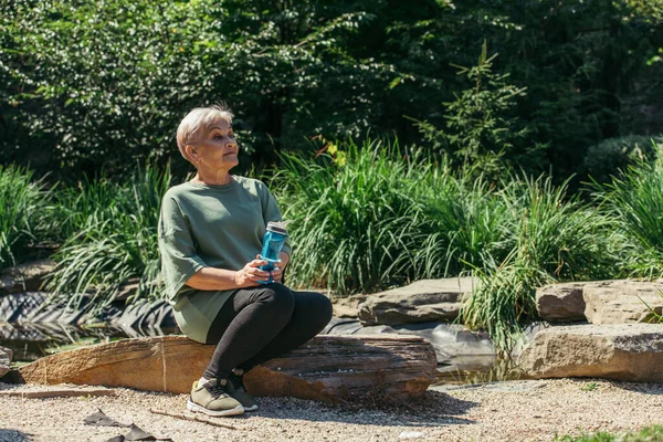 Retired woman in sportswear sitting with sports bottle on wooden log - foto de stock