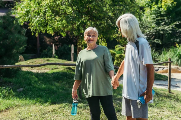 Happy retired couple in sportswear with sport bottles holding hands in park — Foto stock