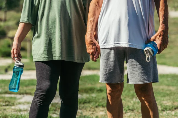 Cropped view of retired couple in sportswear with sport bottles holding hands in park — Stock Photo