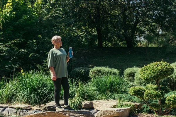 Full length of retired woman in sportswear sitting with sports bottle standing around green plants — Foto stock