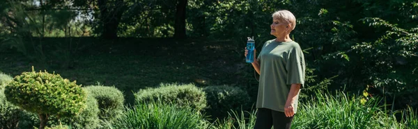 Retired woman in sportswear sitting with sports bottle standing around green plants, banner — Photo de stock