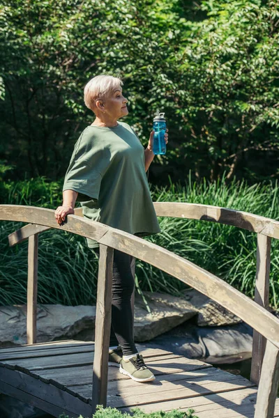 Полная длина пенсионерки в спортивной одежде держа спортивную бутылку и стоя на мосту — стоковое фото