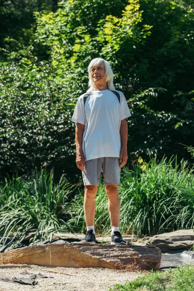 Полная длина пожилого человека с седыми волосами, стоящими в спортивной одежде в зеленом парке — стоковое фото