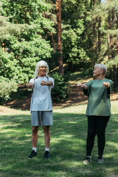Full length of joyful senior couple in sportswear exercising with outstretched hands together in park — Stock Photo
