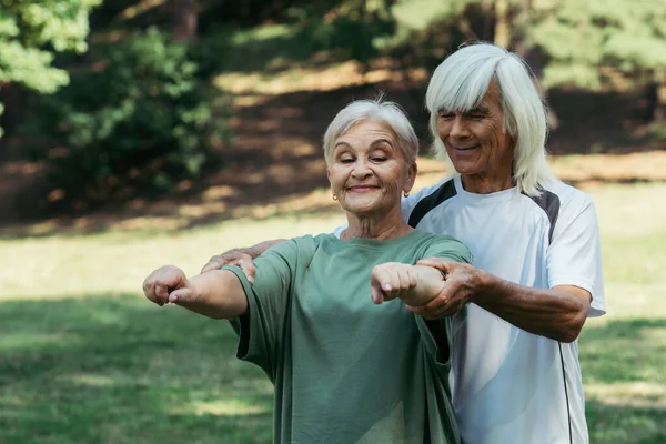 Cheerful senior man in sportswear training happy wife with grey hair in park - foto de stock