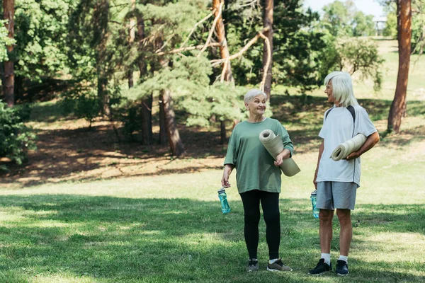 Full length of cheerful senior couple in sportswear holding fitness mats and sports bottles in park — Fotografia de Stock