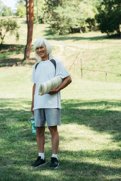 Полная длина счастливого пожилого человека в белой футболке держа спортивную бутылку и фитнес мат в парке — стоковое фото