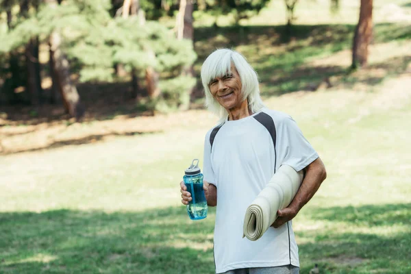 Happy senior man in white t-shirt holding sports bottle and fitness mat in park — Stock Photo