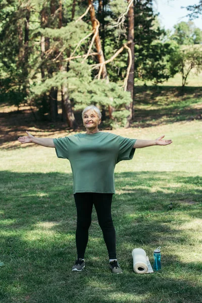 Full length of happy senior woman exercising near sports bottle and fitness mat in park - foto de stock