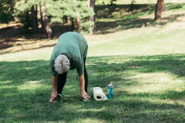 Пенсионерка в полный рост растягивается у спортивной бутылки и фитнес-коврика в парке — стоковое фото