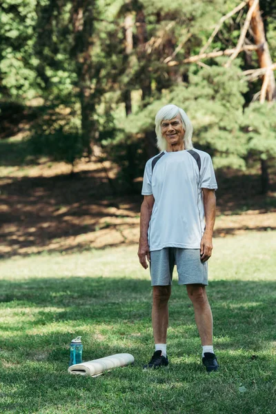 Full length of happy senior man in sportswear standing near sports bottle and fitness mat on grass - foto de stock