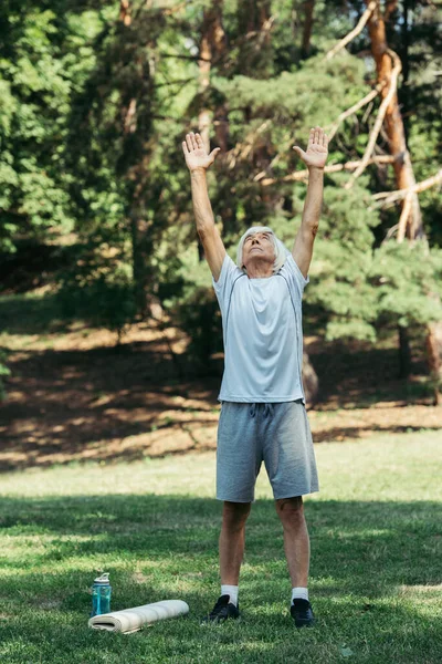 Full length of happy senior man in sportswear exercising near sports bottle and fitness mat on grass — Stock Photo