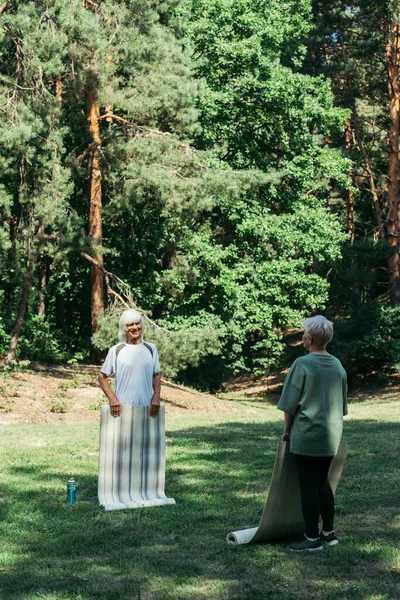 Full length of happy senior man with grey hair looking at wife while holding fitness mat in park — Fotografia de Stock