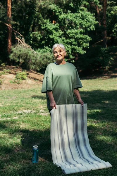 Полная длина счастливой пожилой женщины с седыми волосами, держащей фитнес-коврик в парке — стоковое фото