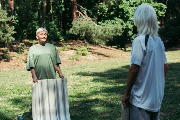 Счастливая пожилая женщина с седыми волосами держа фитнес мат и глядя на мужа в парке — стоковое фото