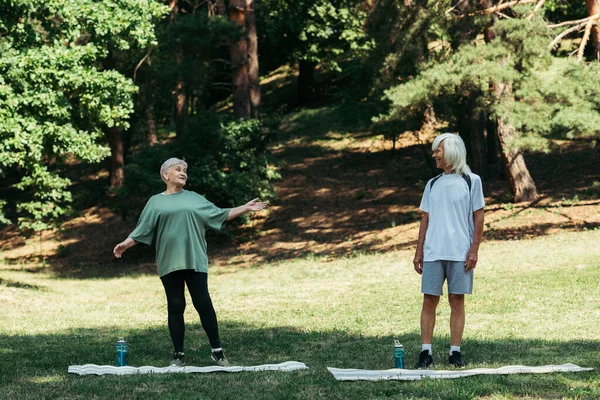 Full length of senior woman pointing with hand at husband with grey hair near fitness mats in park — Stockfoto