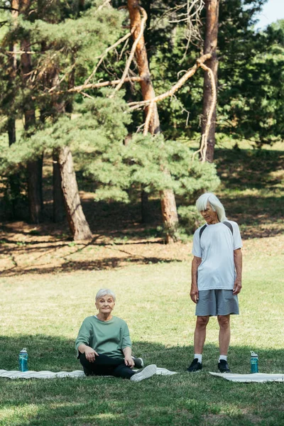 Senior woman sitting on fitness mat near husband with grey hair in park — Stockfoto