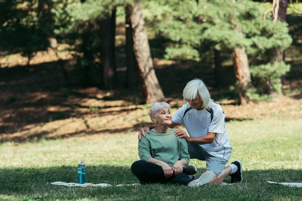 Cheerful senior man hugging wife with grey hair sitting on fitness mat in green park - foto de stock