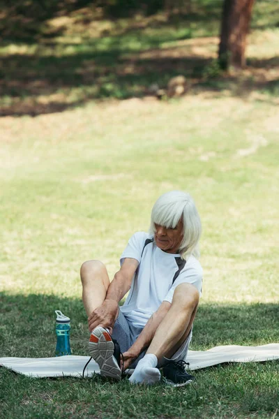 Senior man with grey hair taking off sneakers while sitting on fitness mat in park — Fotografia de Stock