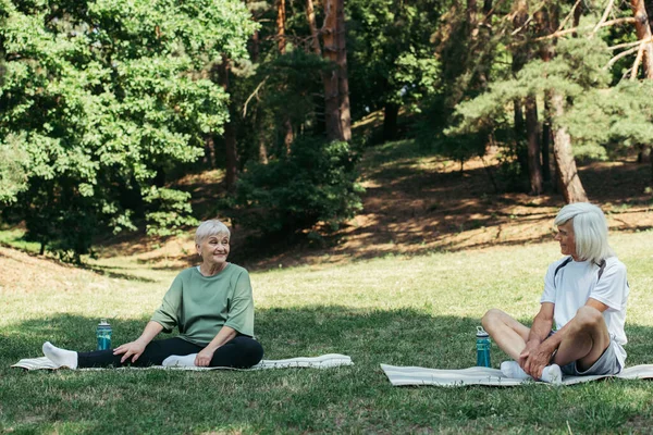 Senior and happy couple sitting on fitness mats and looking at each other in green park — Fotografia de Stock