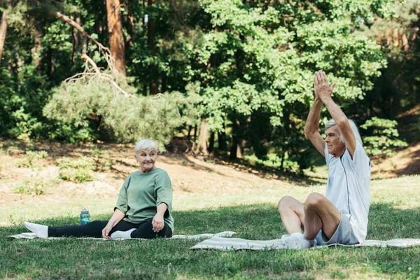 Smiling senior woman looking at husband with praying hands above head practicing on fitness mat in green park — Stock Photo