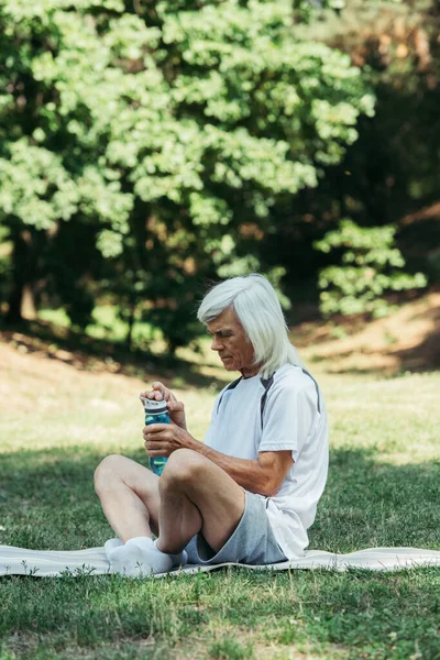 Старший чоловік з сірим волоссям тримає спортивну пляшку, сидячи на фітнес-маті в зеленому парку — стокове фото