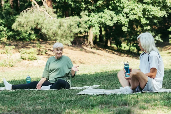 Cheerful senior woman sitting on fitness mat and pointing at husband with sports bottle - foto de stock