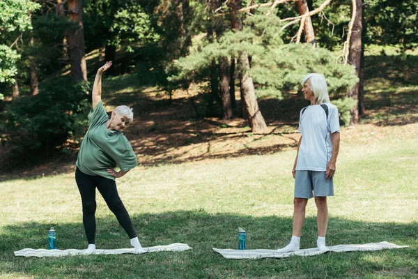 Полный рост пожилой мужчина смотрит на жену с рукой на бедре, тренируясь на фитнес-коврике — стоковое фото