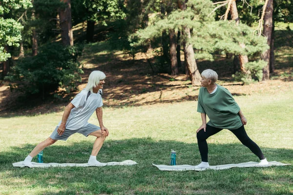 Full length of senior couple with grey hair smiling and doing lunges on fitness mats in park — Stockfoto